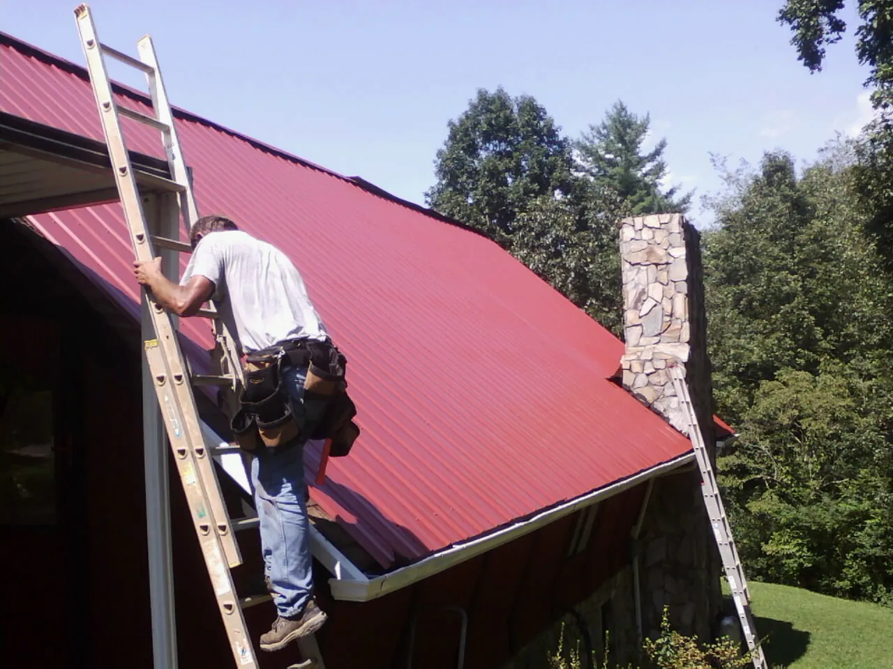 Roofer on a ladder beside a red metal roof and stone chimney — Soffit & Fascia Repair by Roofing Brothers in Penn Yan